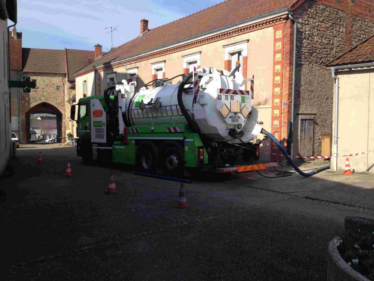 Camion de ramassage vert et blanc stationné dans une rue pavée, tuyau d'arrosage raccordé à une bouche d'égout.