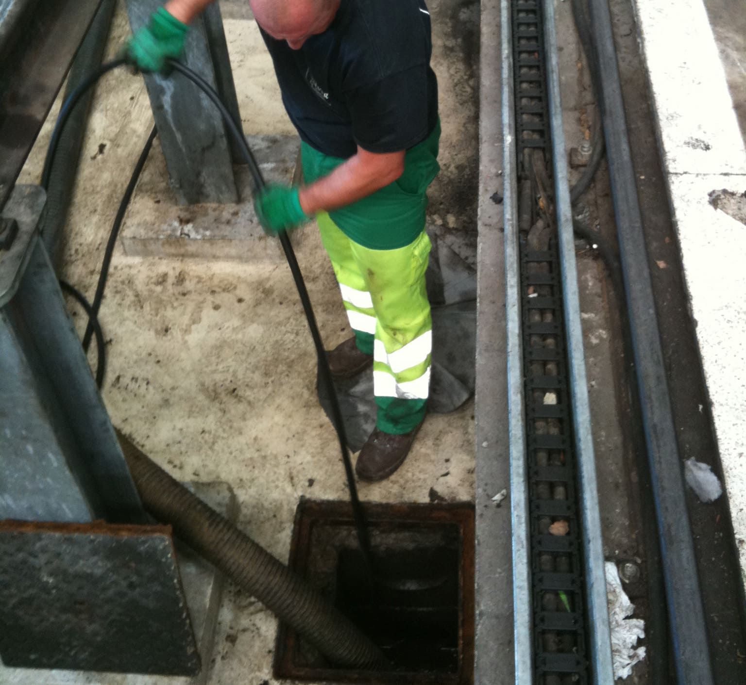 Un homme en tenue de travail nettoie une canalisation avec un tuyau d'arrosage à l'extérieur.