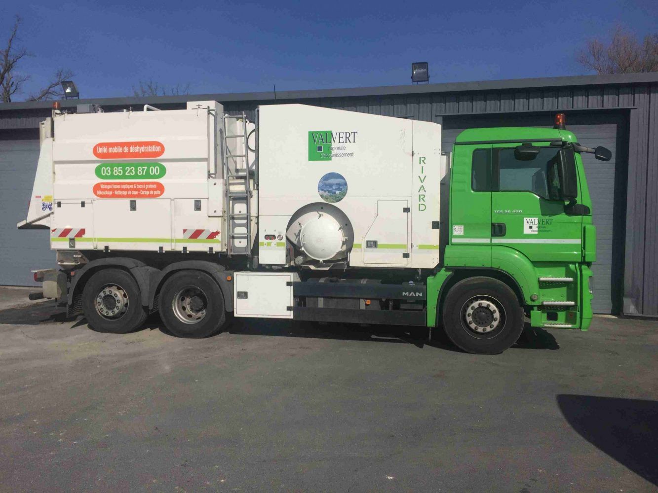 Camion-poubelle vert et blanc stationné devant un bâtiment gris par une journée ensoleillée.