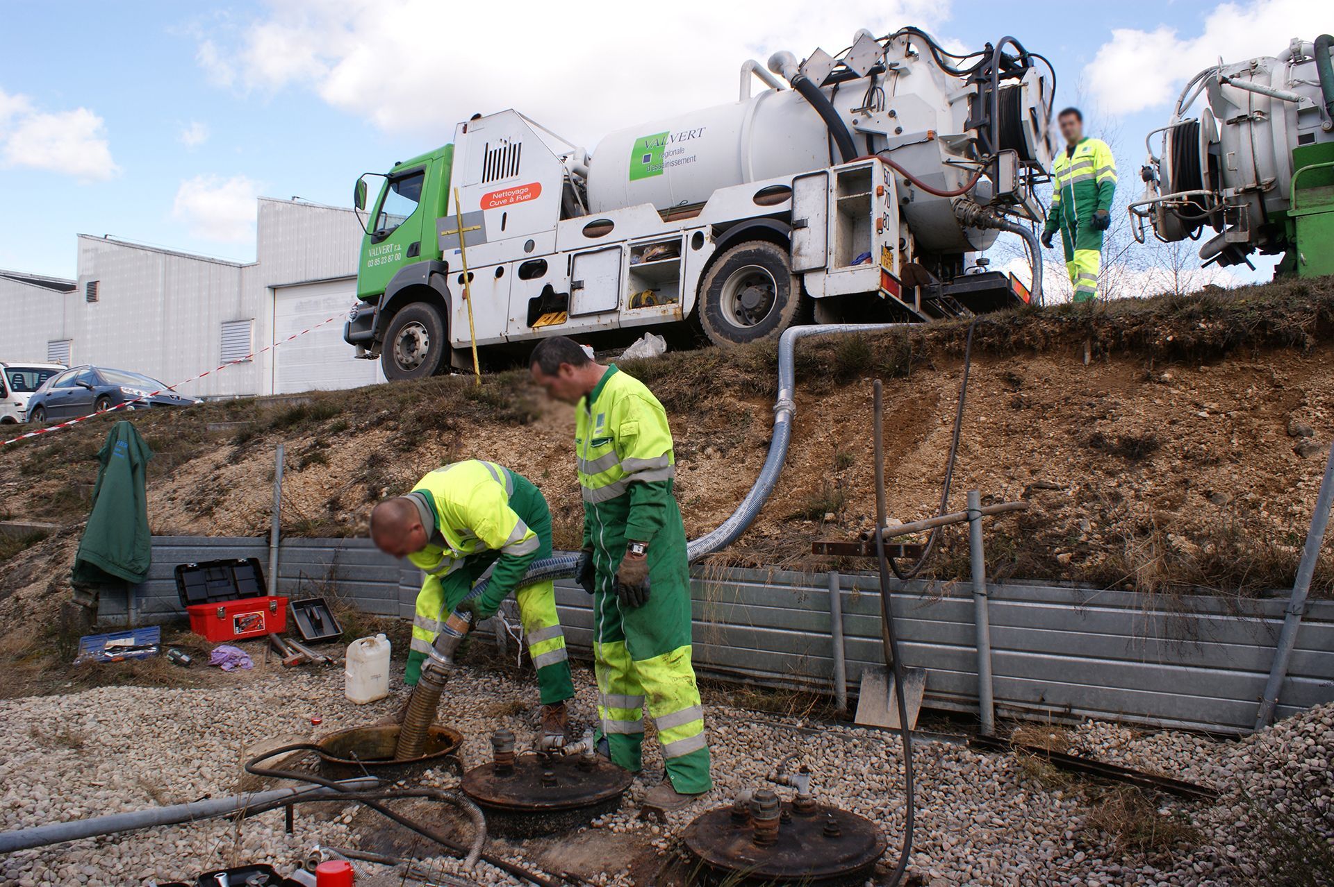 Des ouvriers portant des gilets réfléchissants et un camion équipé d'un tuyau d'aspiration nettoient un système d'égouts à flanc de colline.