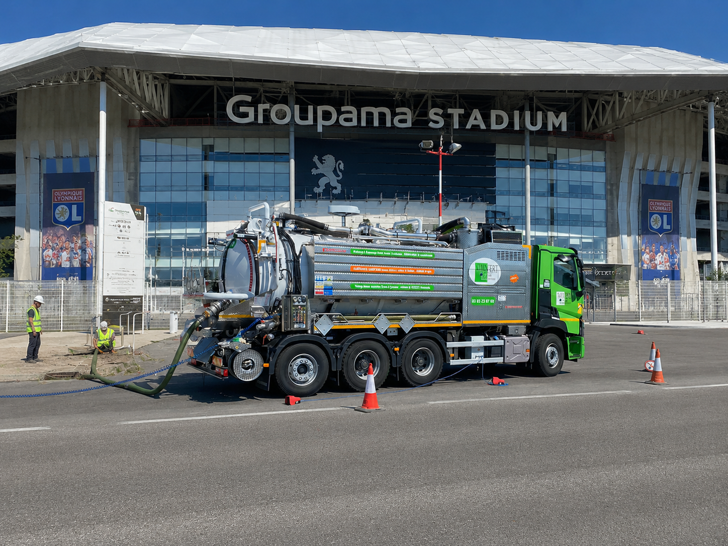 Camion de ramassage vert et blanc stationné dans une rue pavée, tuyau d'arrosage raccordé à une bouche d'égout.