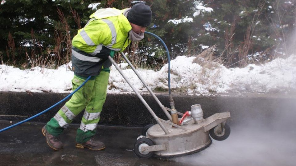 Homme en combinaison vert fluo nettoyant une surface asphaltée au nettoyeur haute pression ; fond enneigé.