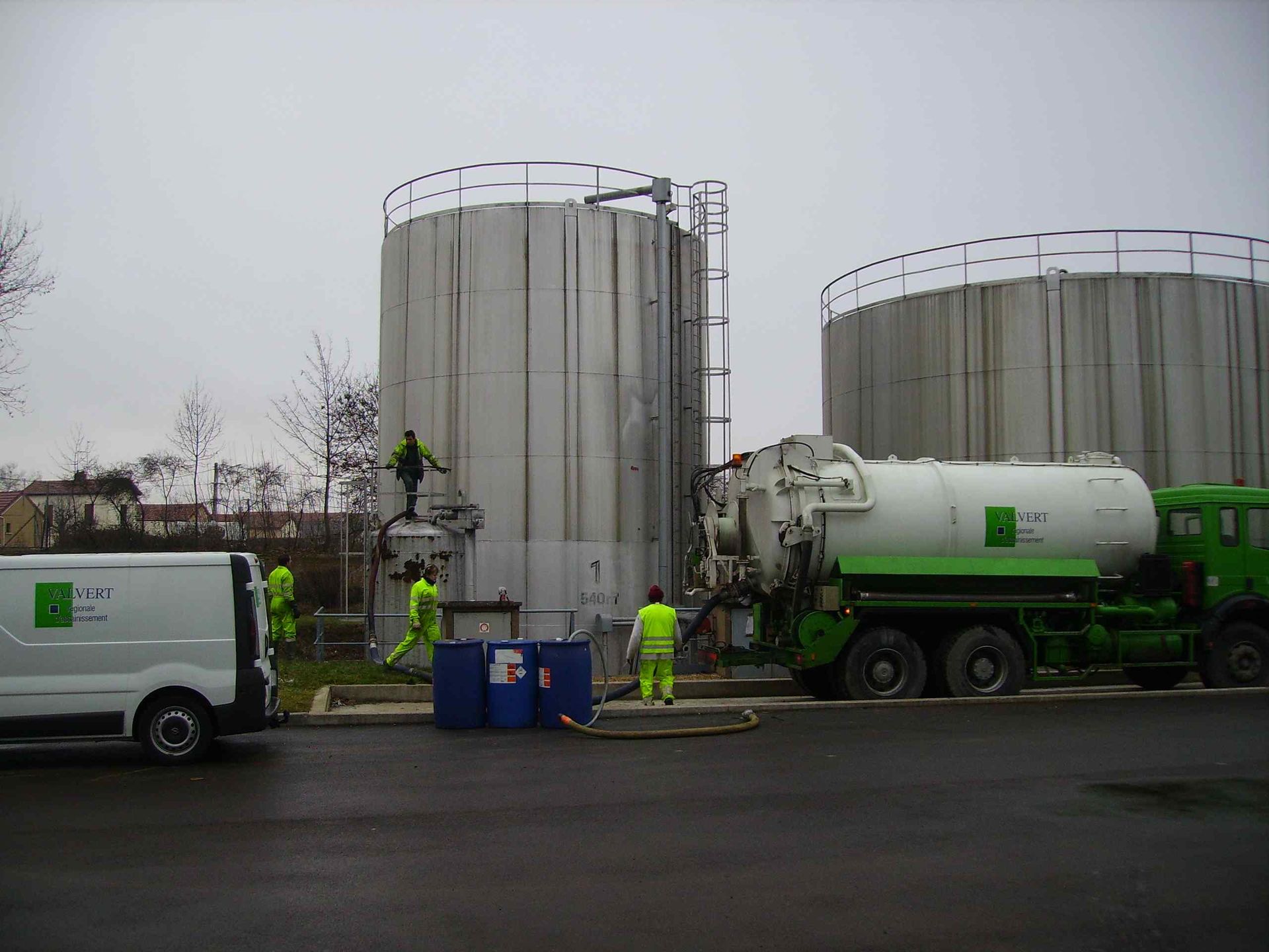 Des ouvriers portant des gilets de sécurité se trouvent près de réservoirs industriels et d'un camion-citerne vert, probablement chargé du transport de déchets liquides.