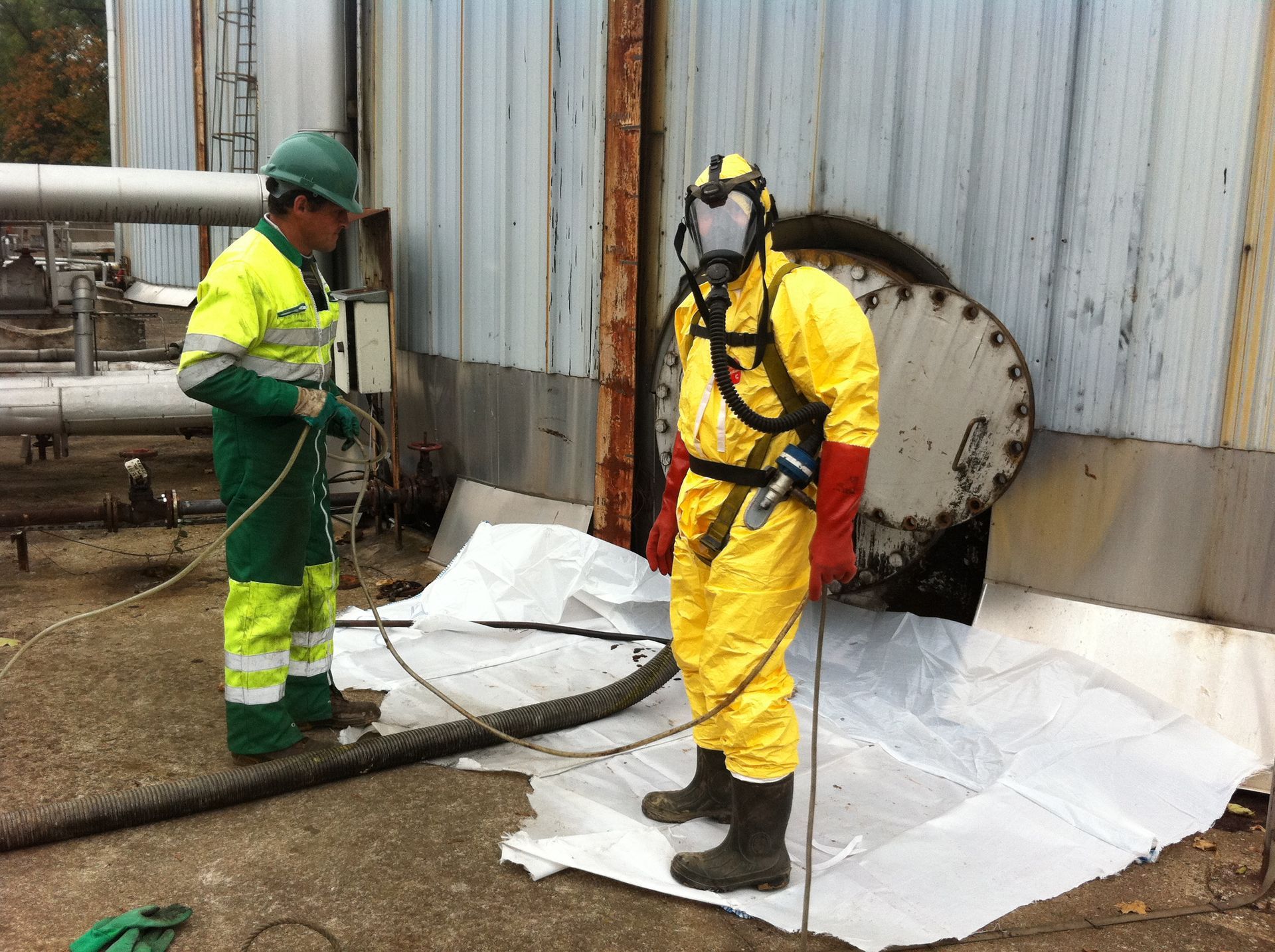 Deux ouvriers : l'un en combinaison de protection chimique près de l'ouverture du réservoir, l'autre en vêtements de travail. Environnement extérieur.