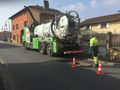 Un camion-citerne vert et blanc aspire une plaque d'égout sur une rue de la ville ; un ouvrier en tenue de sécurité se tient à proximité.