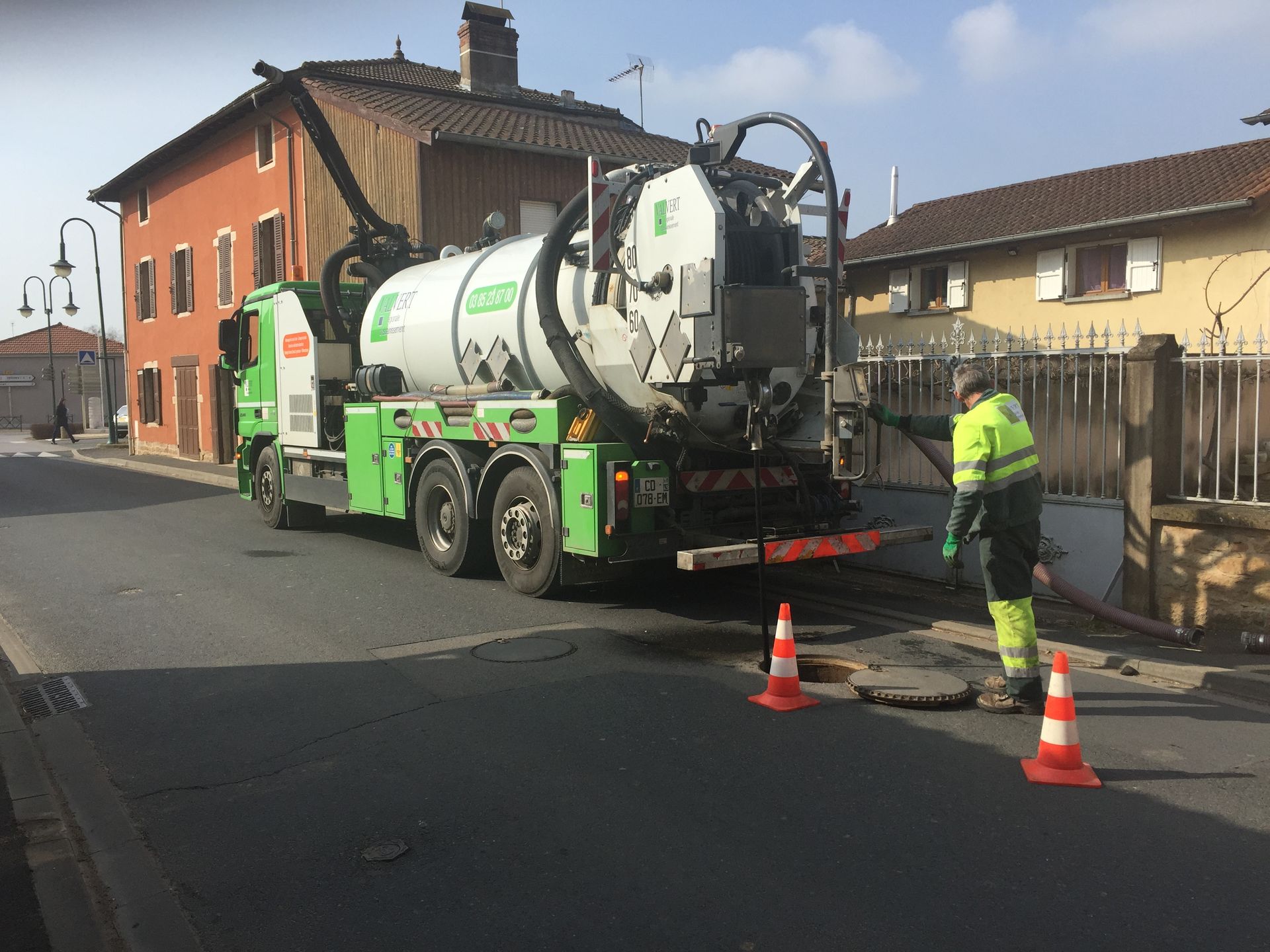 Camion de ramassage vert stationné dans la rue, employé en gilet de sécurité près d'une bouche d'égout ouverte.