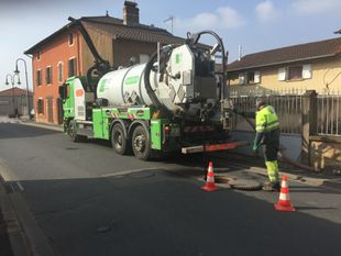 Un camion-citerne vert et blanc aspire une plaque d'égout sur une rue de la ville ; un ouvrier en tenue de sécurité se tient à proximité.
