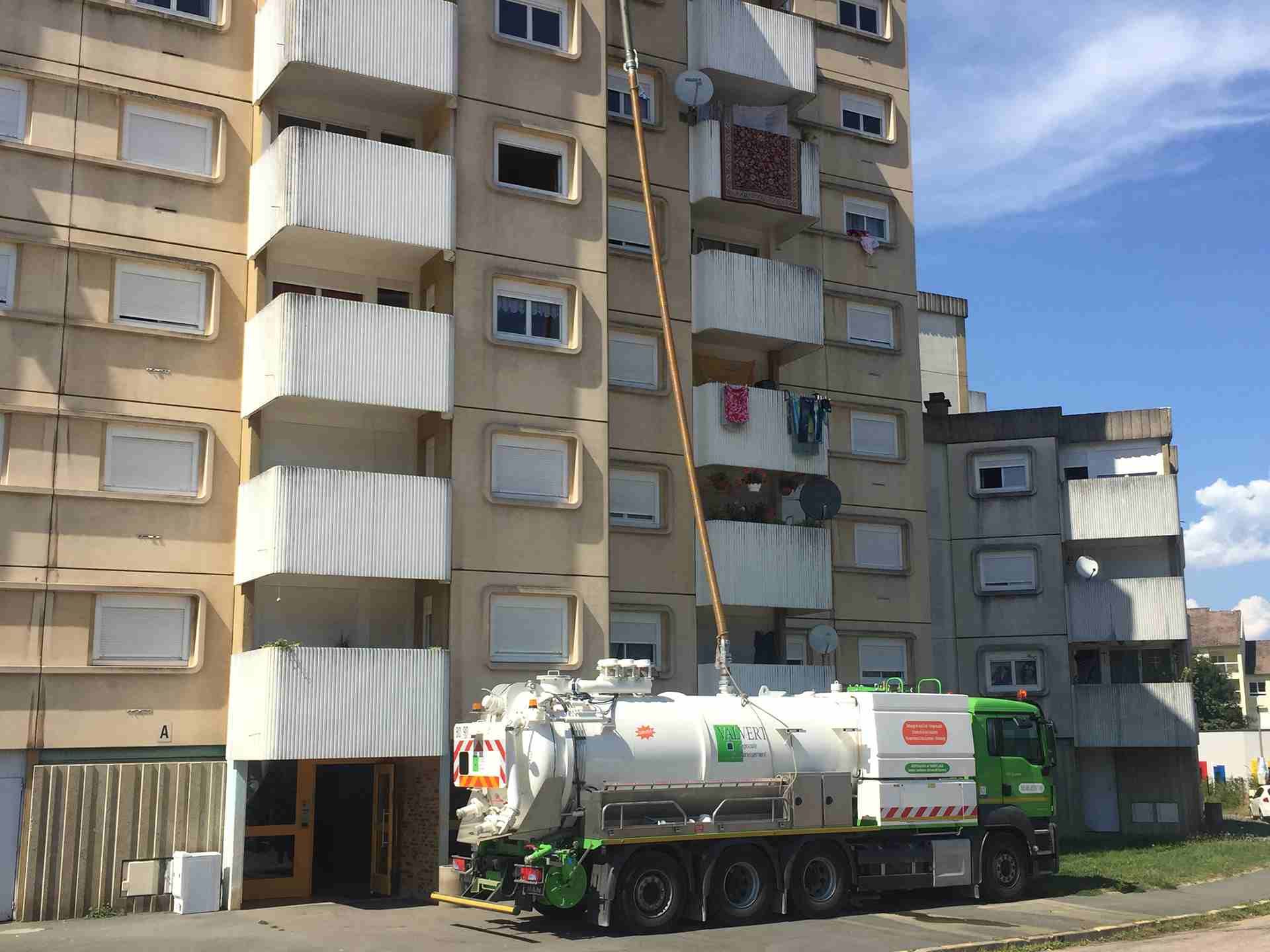 Camion avec un long tuyau à côté d'un immeuble d'appartements, probablement pour le nettoyage ou l'entretien.
