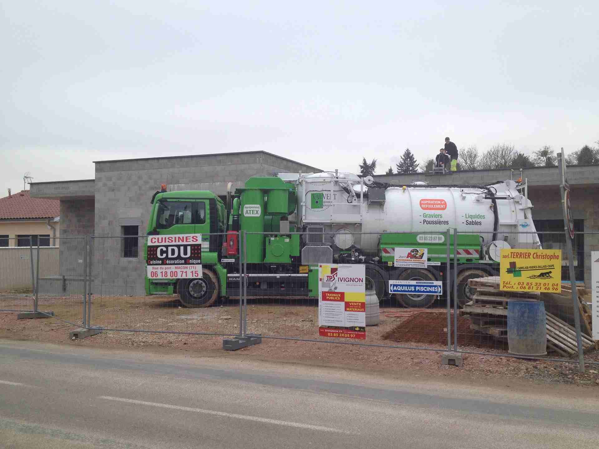 Un camion industriel vert et blanc est stationné près d'un bâtiment en construction. Une personne se trouve sur le toit.