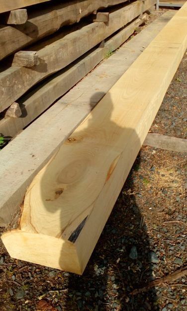Primer plano de una viga de madera larga y de color claro con una muesca tallada, apilada junto a otras piezas de madera al aire libre.