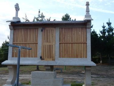 Pequeña estructura de madera elevada con una puerta central, sobre bloques de hormigón al aire libre, con un brazo de metal azul cerca.