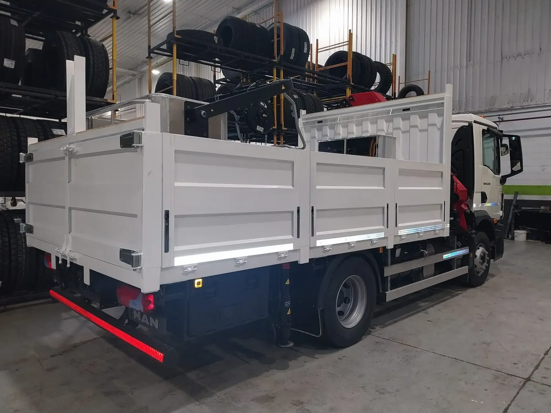 Camión blanco de plataforma, estacionado en el interior, cargado de neumáticos. Bandas reflectantes visibles.