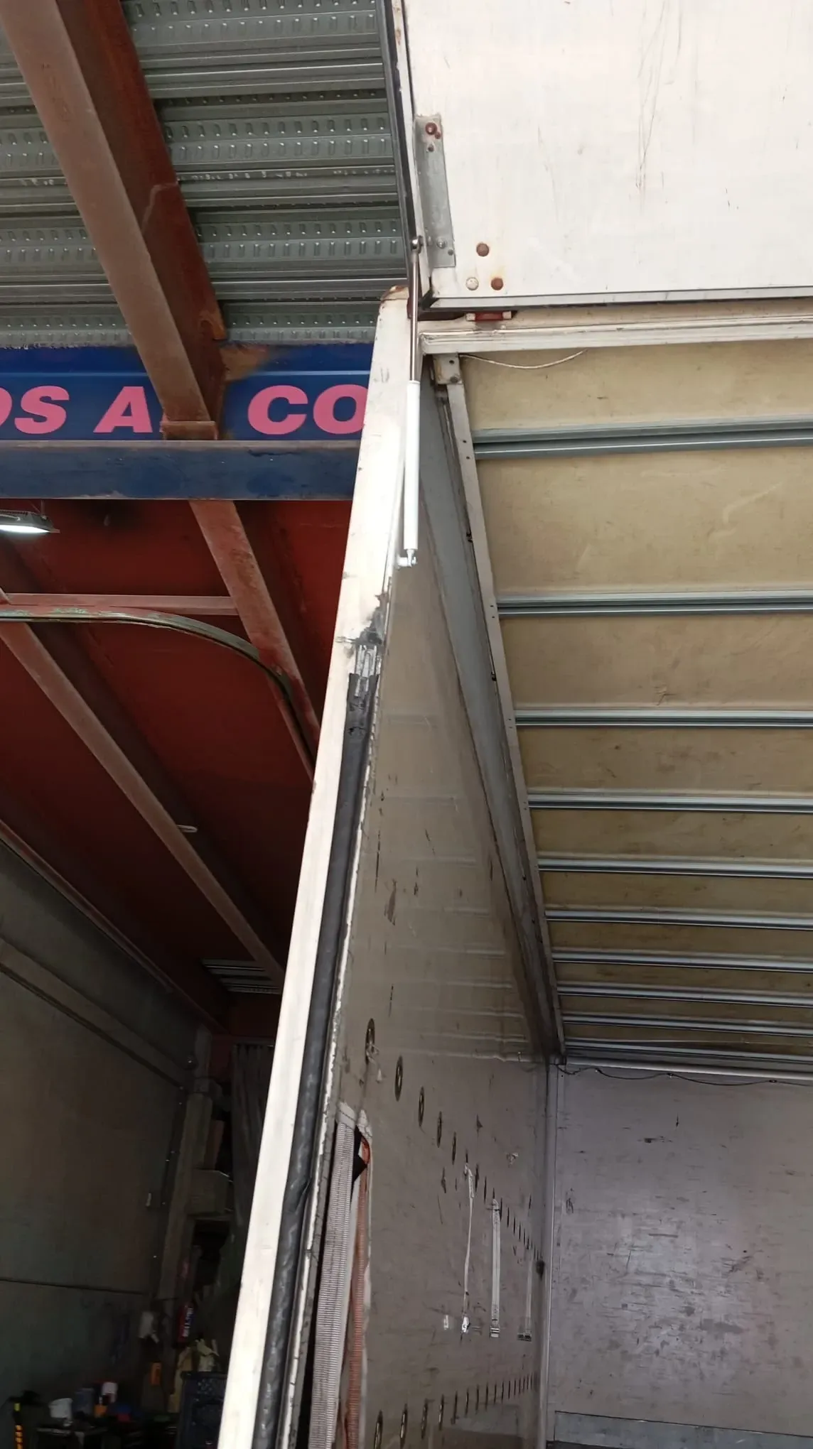 Vista lateral del área de carga de un camión parcialmente abierta. El lateral metálico presenta daños y se aprecia parcialmente una señal roja y azul.