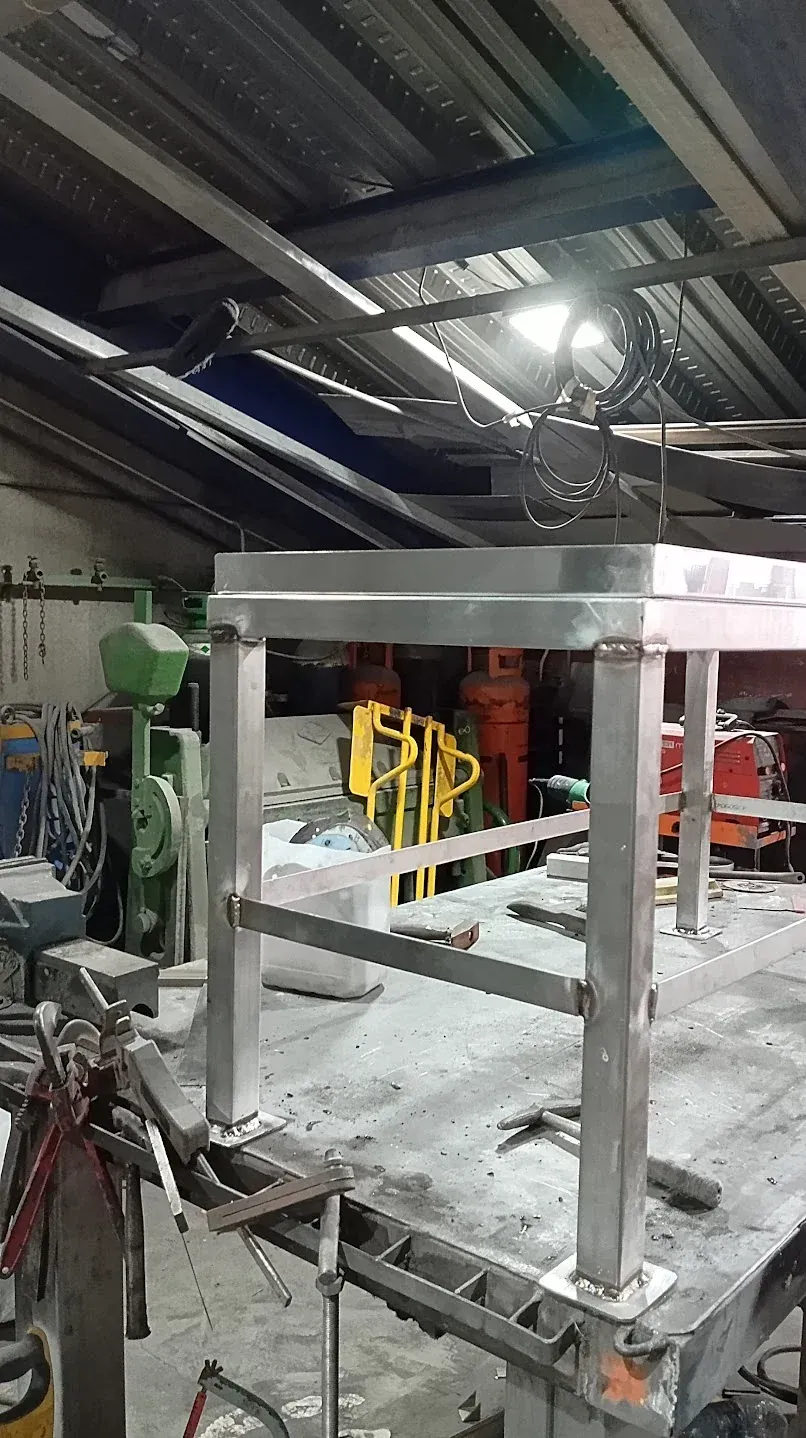 Trabajos de metal en proceso en un taller. Se destaca una estructura de mesa parcialmente construida, con otras máquinas al fondo.