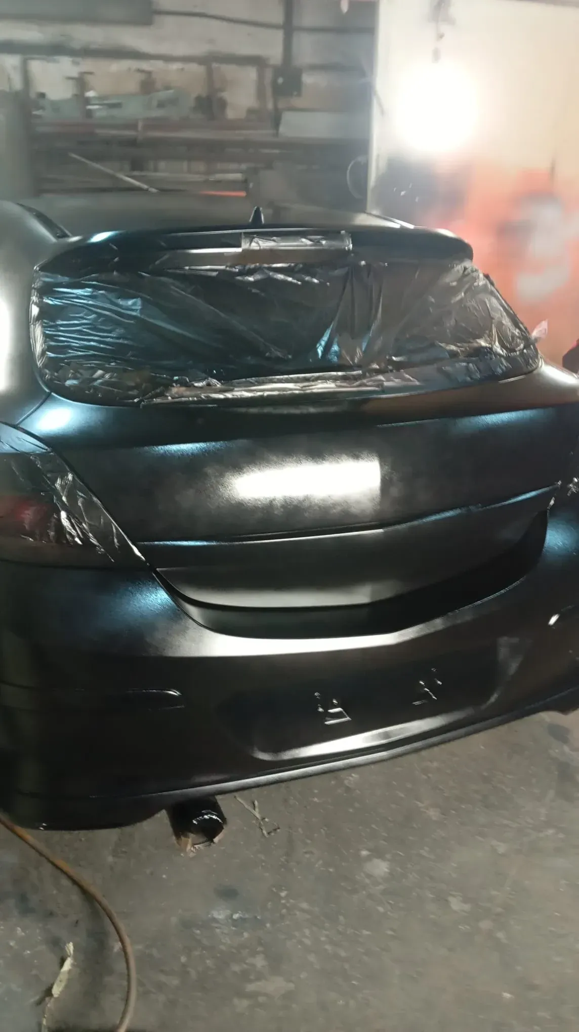 Coche negro siendo pintado en taller, parte trasera recubierta de plástico, en negro mate.