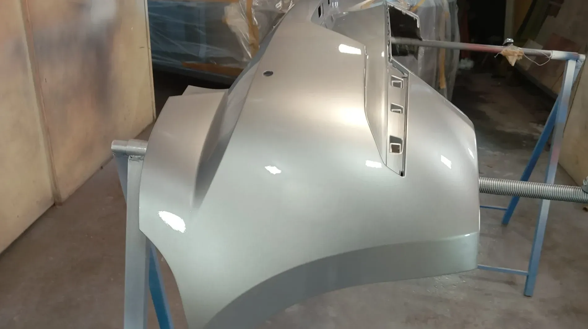 Parachoques de coche plateado, recién pintado, apoyado sobre un soporte en un taller.