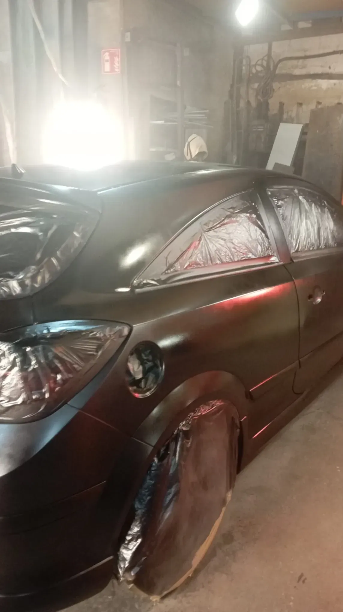 Coche pintado de negro en un garaje; el rojo es visible a través de las ventanas cubiertas con cinta adhesiva, las partes protegidas están cubiertas con plástico.