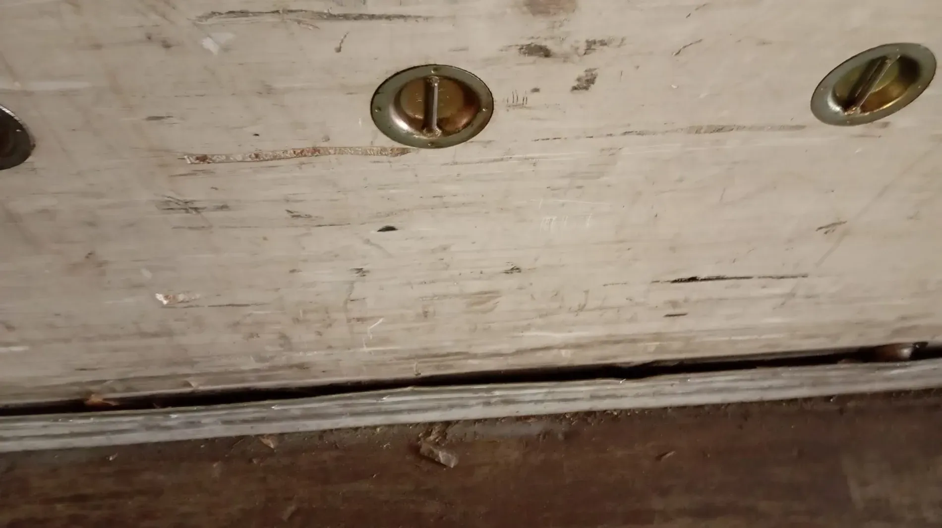 Primer plano de una superficie de madera blanca rayada con tres tornillos visibles en un metal incrustado de color dorado.