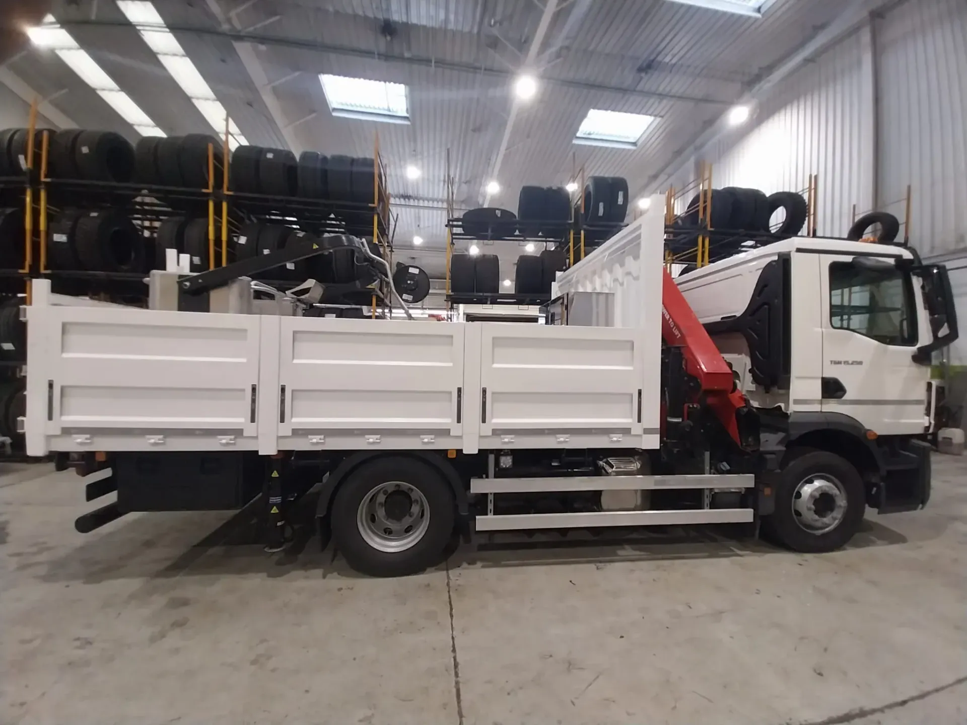 Camión de plataforma blanco con grúa en un almacén, neumáticos en estantes al fondo.