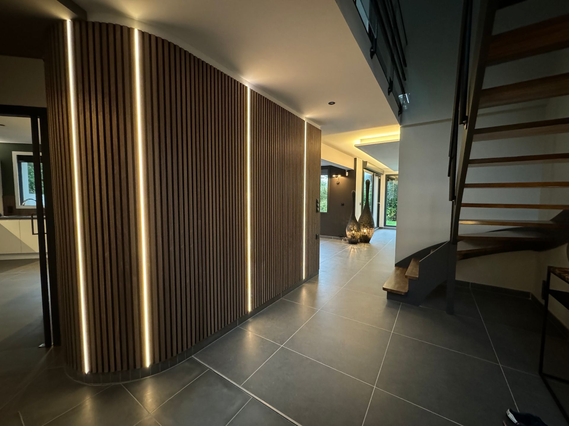 Couloir moderne avec murs lambrissés en bois foncé, sol carrelé, escalier à droite et éclairage chaleureux