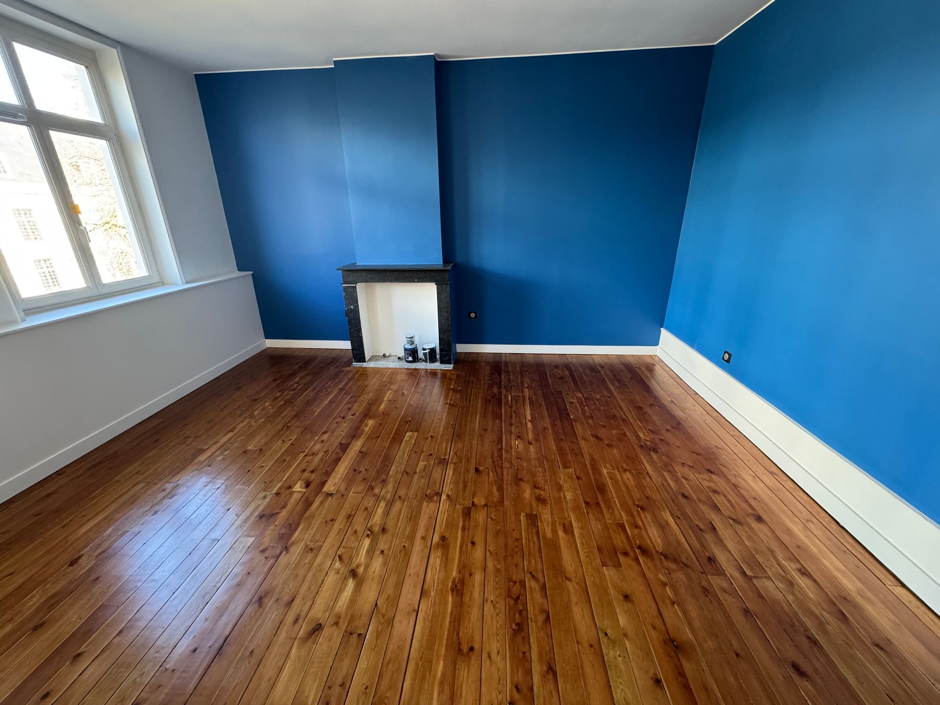 pièce avec fenêtre ancienne sur la gauche, cheminée dans le mur du fond, murs peints en bleu, parquet brillant