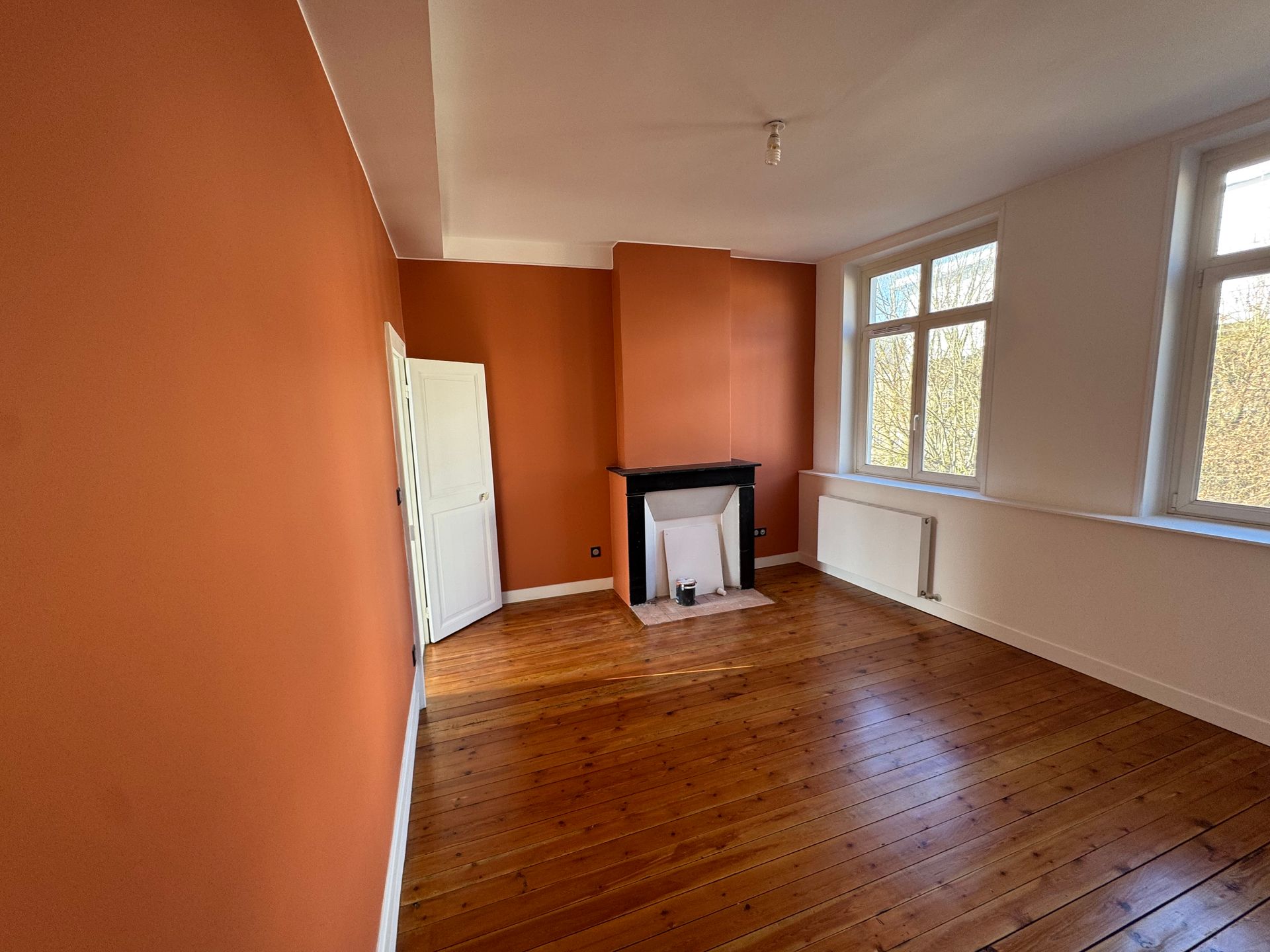 Pièce vide aux murs orange, parquet, cheminée blanche et grandes fenêtres sur un côté