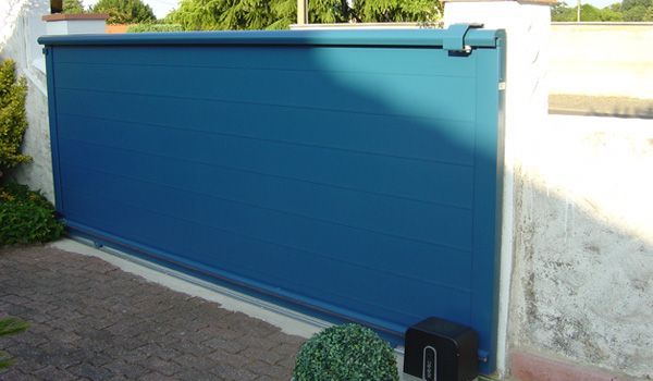 Portail roulant bleu, fermant l'entrée d'une propriété, installé sur des piliers blancs, dans un cadre extérieur ensoleillé.