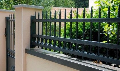 Clôture en métal noir ornée de cercles et de pointes décoratives, surmontant un mur beige à côté d'un portail.