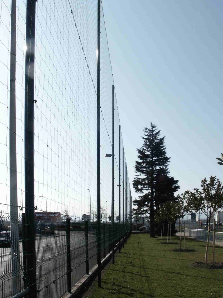 Une clôture en grillage borde une zone herbeuse arborée, sous un ciel d'un bleu éclatant.