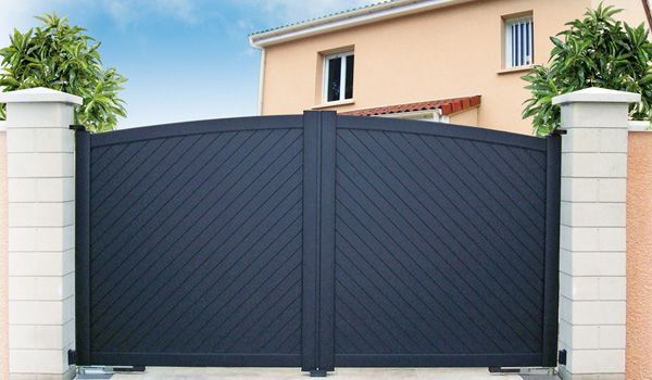 Portail double noir à motif diagonal, à sommet arqué, entre deux piliers de briques à coiffe blanche.