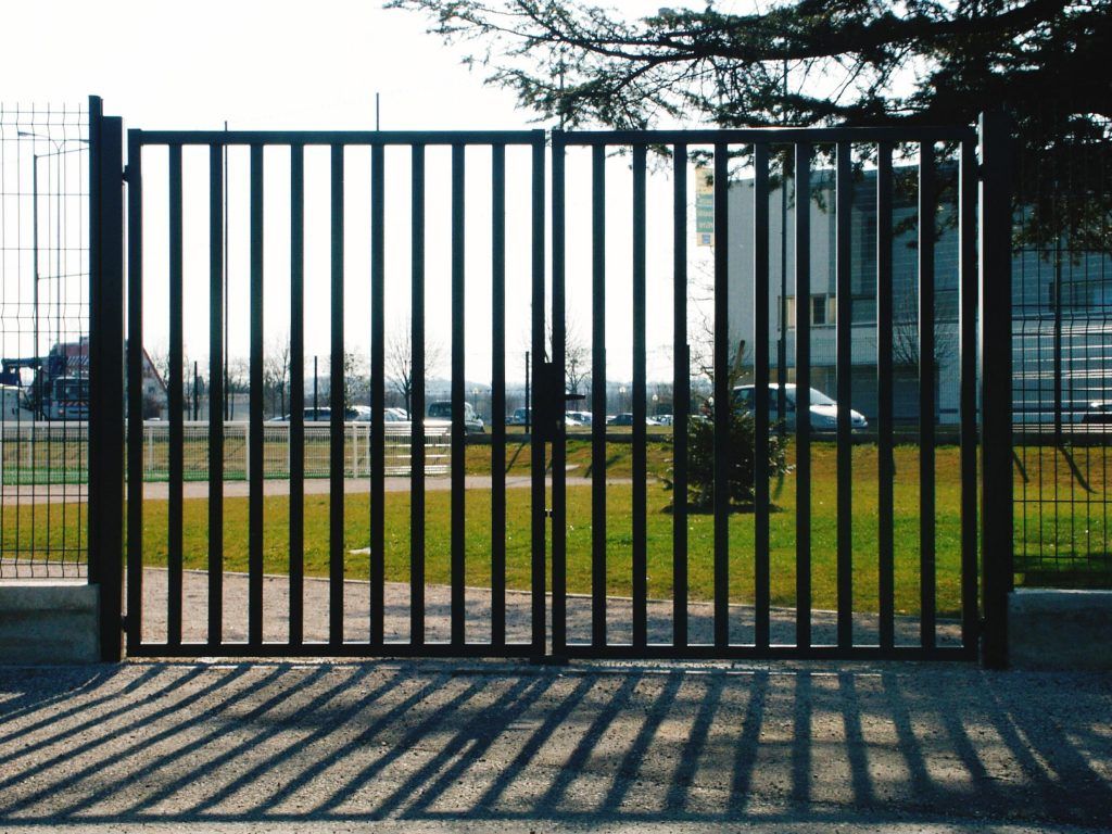 Un portail en métal noir à barreaux verticaux projette une ombre sur le sol. À l'arrière-plan, on aperçoit de l'herbe verte et des bâtiments.