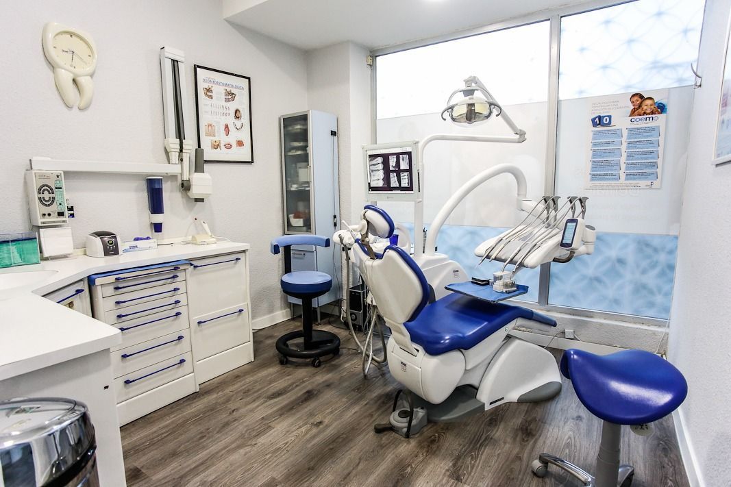 Interior de consultorio dental con silla azul y blanca, armarios y equipo. Una ventana proporciona luz natural.