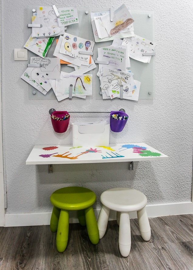 Estación de arte infantil con una mesa blanca y taburetes bajo un tablero de vidrio que exhibe obras de arte.