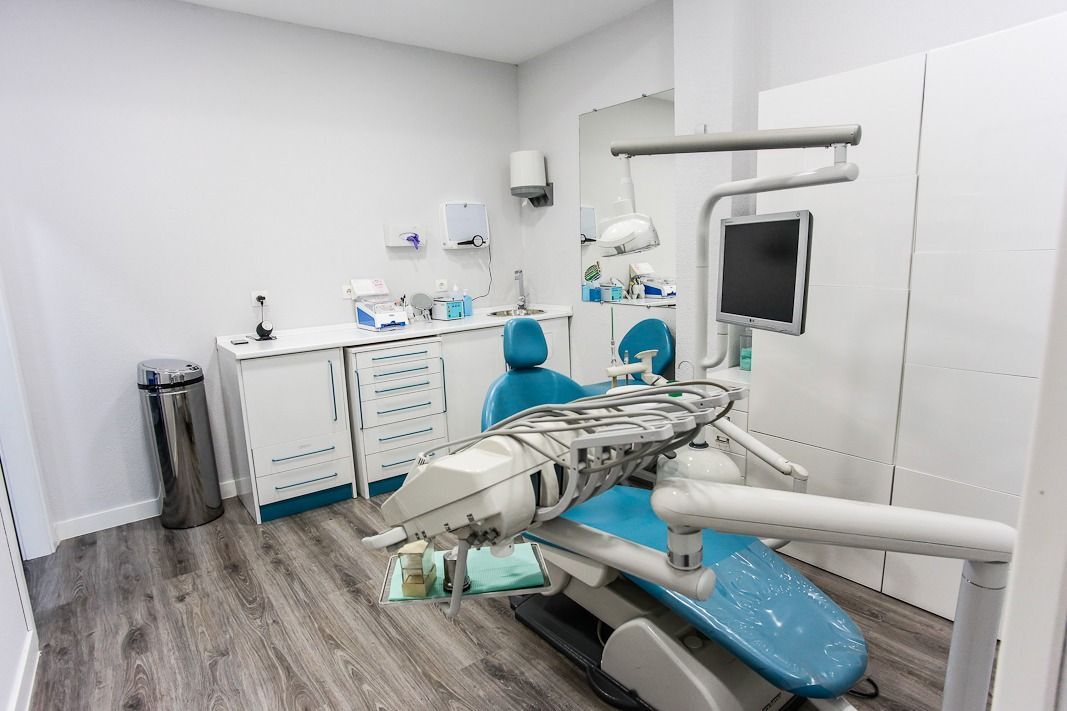 Consultorio dental con sillón azul, armarios blancos y suelo gris. La sala está muy iluminada.