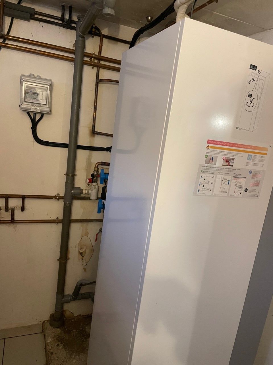 Appareil électroménager blanc à côté de tuyaux et d'un tableau électrique dans un sous-sol.