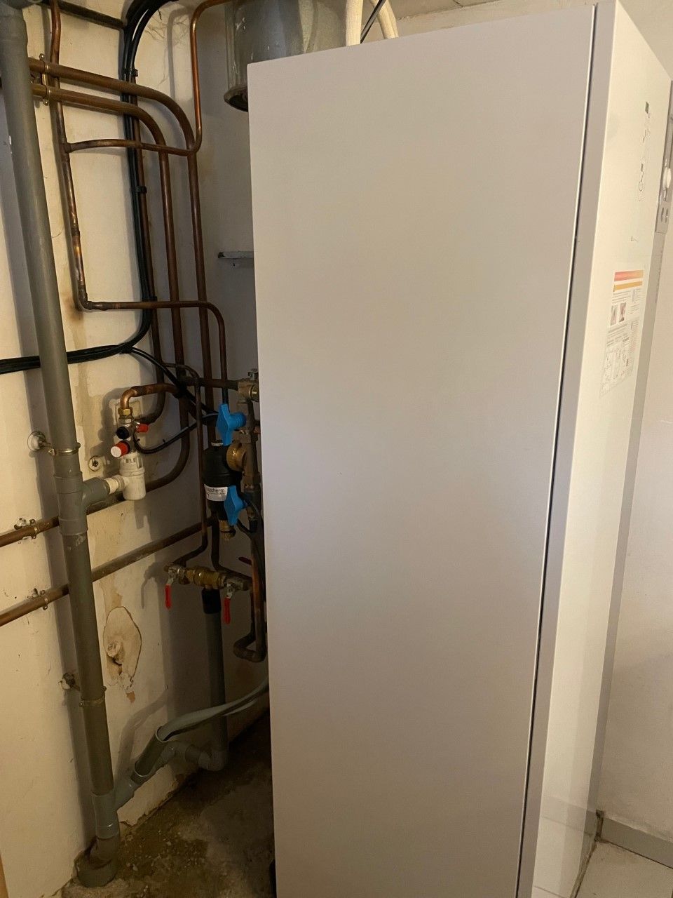 Un chauffe-eau encastré dans un panneau clair, avec des tuyaux et une plomberie en cuivre contre un mur blanc.