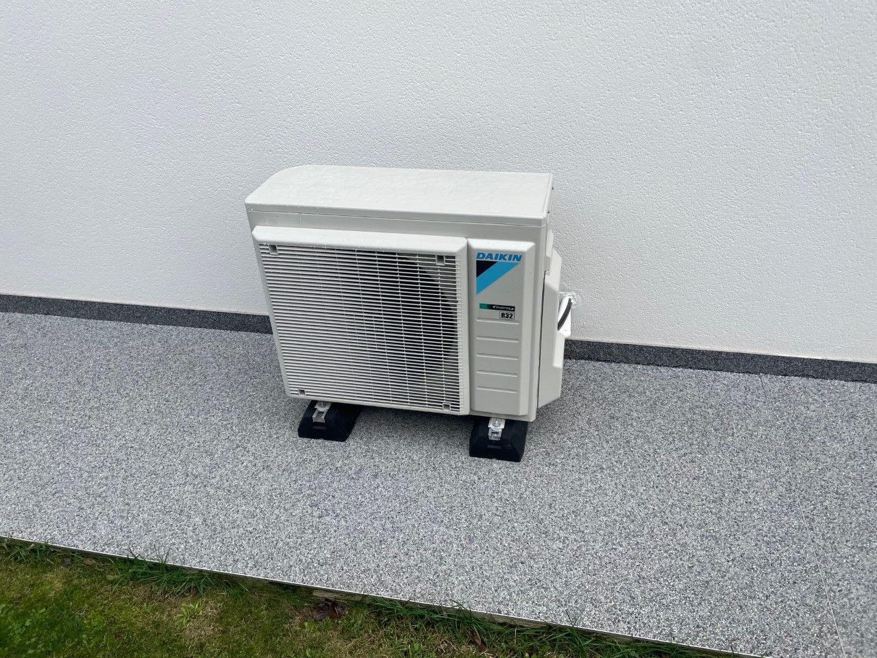 Unité de climatisation Daikin montée sur des blocs noirs contre un mur en stuc blanc et du gravier.