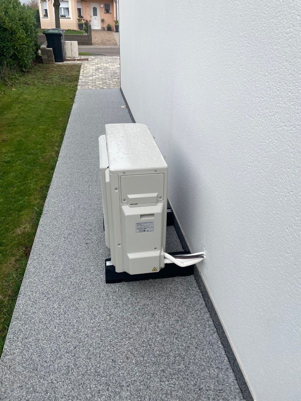 Unité de climatisation sur un support noir à côté d'un mur blanc, sur un chemin de gravier.