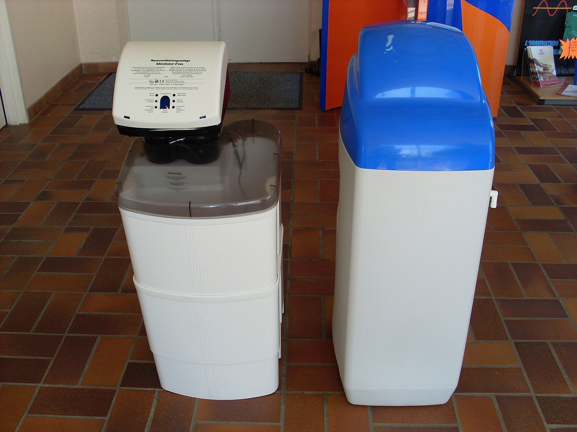 Deux systèmes d'adoucissement d'eau ; l'un avec un panneau de commande, tous deux dans une pièce avec un sol carrelé.