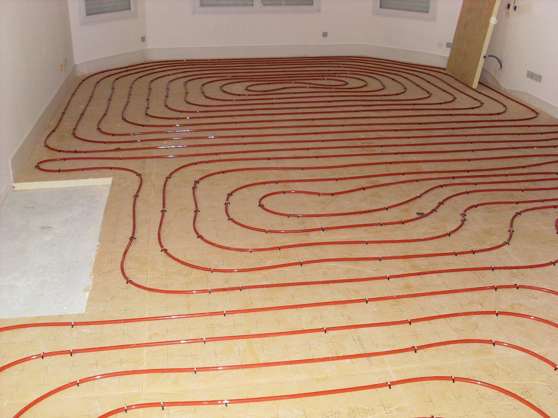 Des tuyaux rouges serpentent sur le sol, probablement pour un chauffage au sol radiant, dans une pièce aux murs blancs.