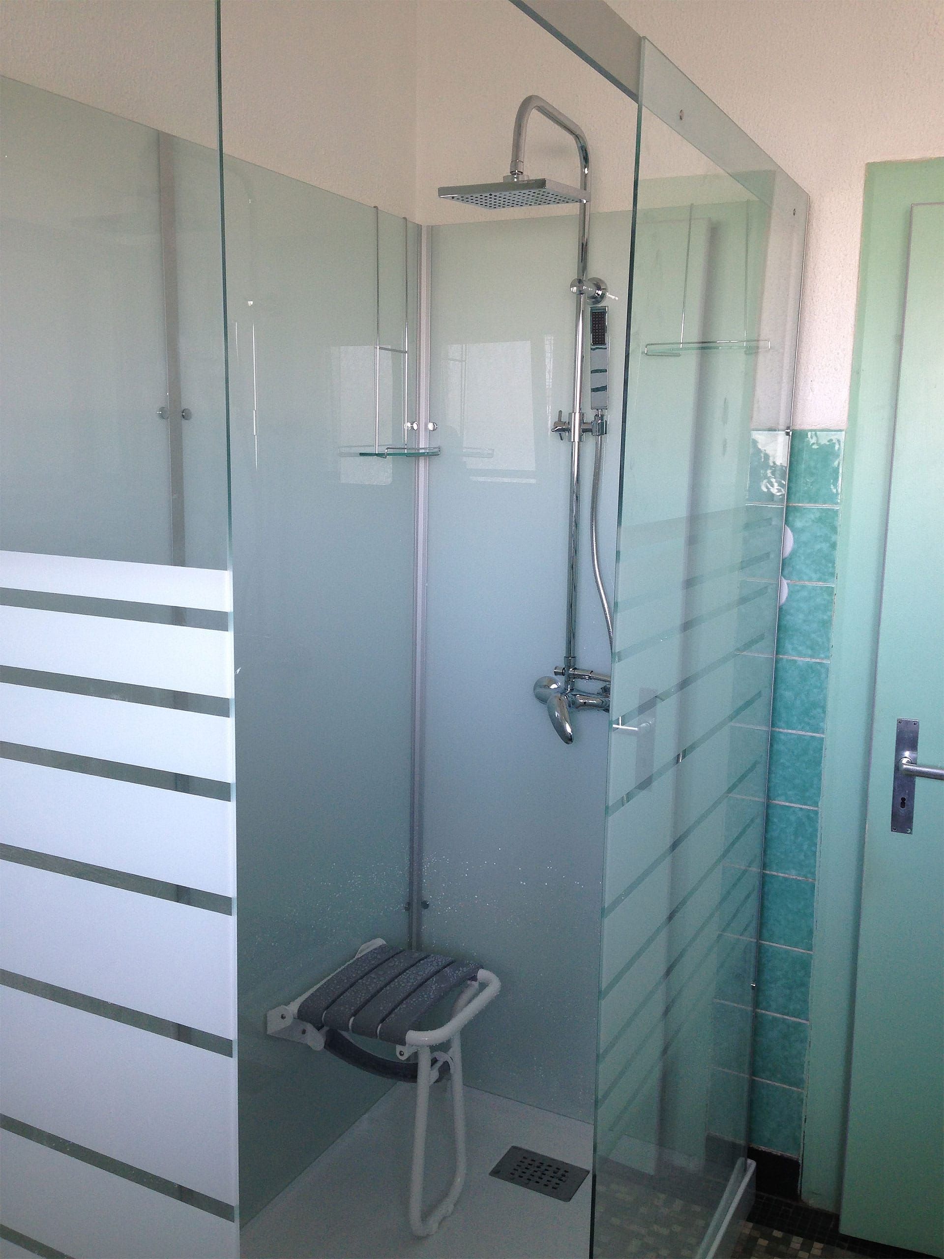 Cabine de douche avec portes en verre, siège de douche et pommeau de douche effet pluie. Les murs sont bleu sarcelle et dépolis.