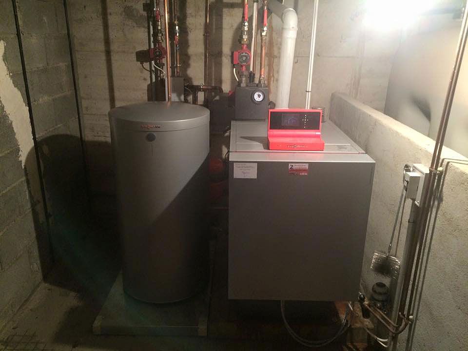 Chauffe-eau à eau grise et chaudière installés au sous-sol, avec des tuyaux en cuivre et un appareil rouge au-dessus.