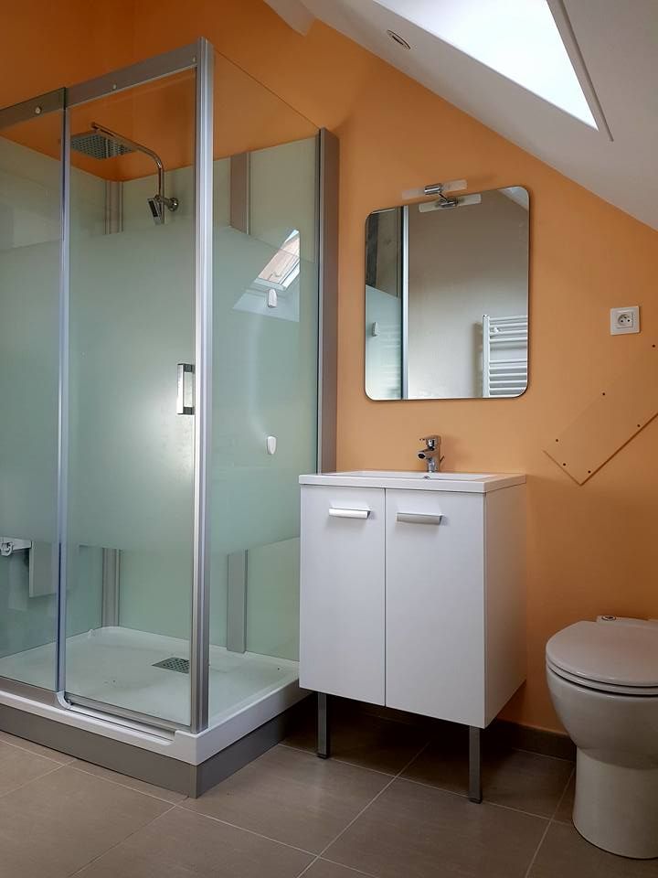 Salle de bains avec cabine de douche, meuble-lavabo, miroir et toilettes, contre un mur orange, plafond en pente.