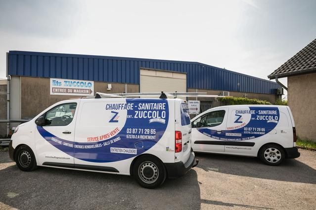 Deux camionnettes de livraison blanches avec des logos bleus sont garées devant un bâtiment industriel bleu.