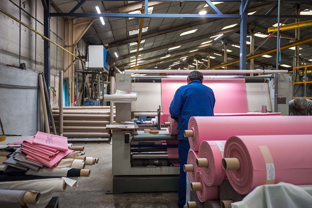 Un ouvrier en salopette bleue actionne une machine ; des rouleaux de tissu rose sont visibles dans une usine.