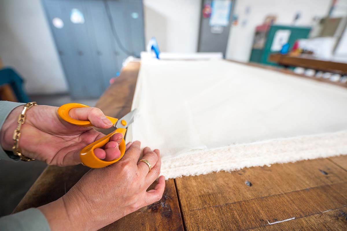 Des mains découpent du tissu blanc avec des ciseaux orange sur une table en bois.