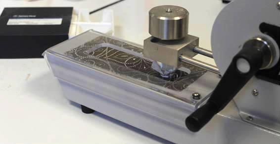 Gros plan sur une machine avec un socle argenté, un cylindre métallique et une manivelle ; une surface blanche est en cours de traitement.