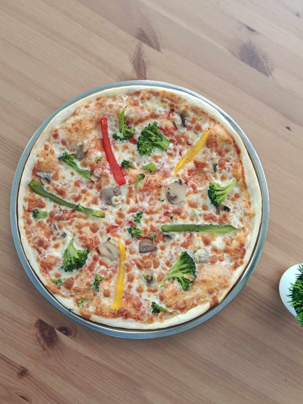 Una pizza vegetariana con brócoli, pimientos y champiñones en una sartén plateada sobre una mesa de madera.