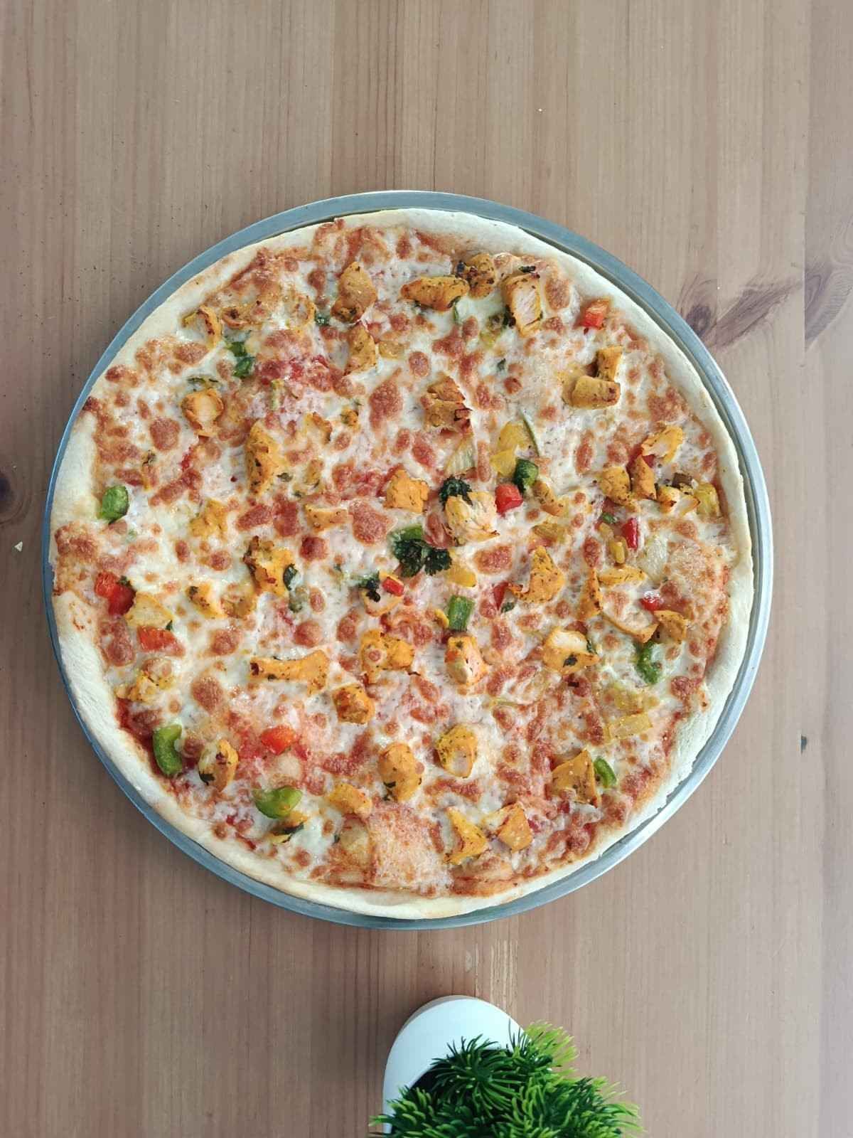 Una pizza redonda de pollo con queso fundido y pimientos, servida en una bandeja de metal sobre una mesa de madera clara.