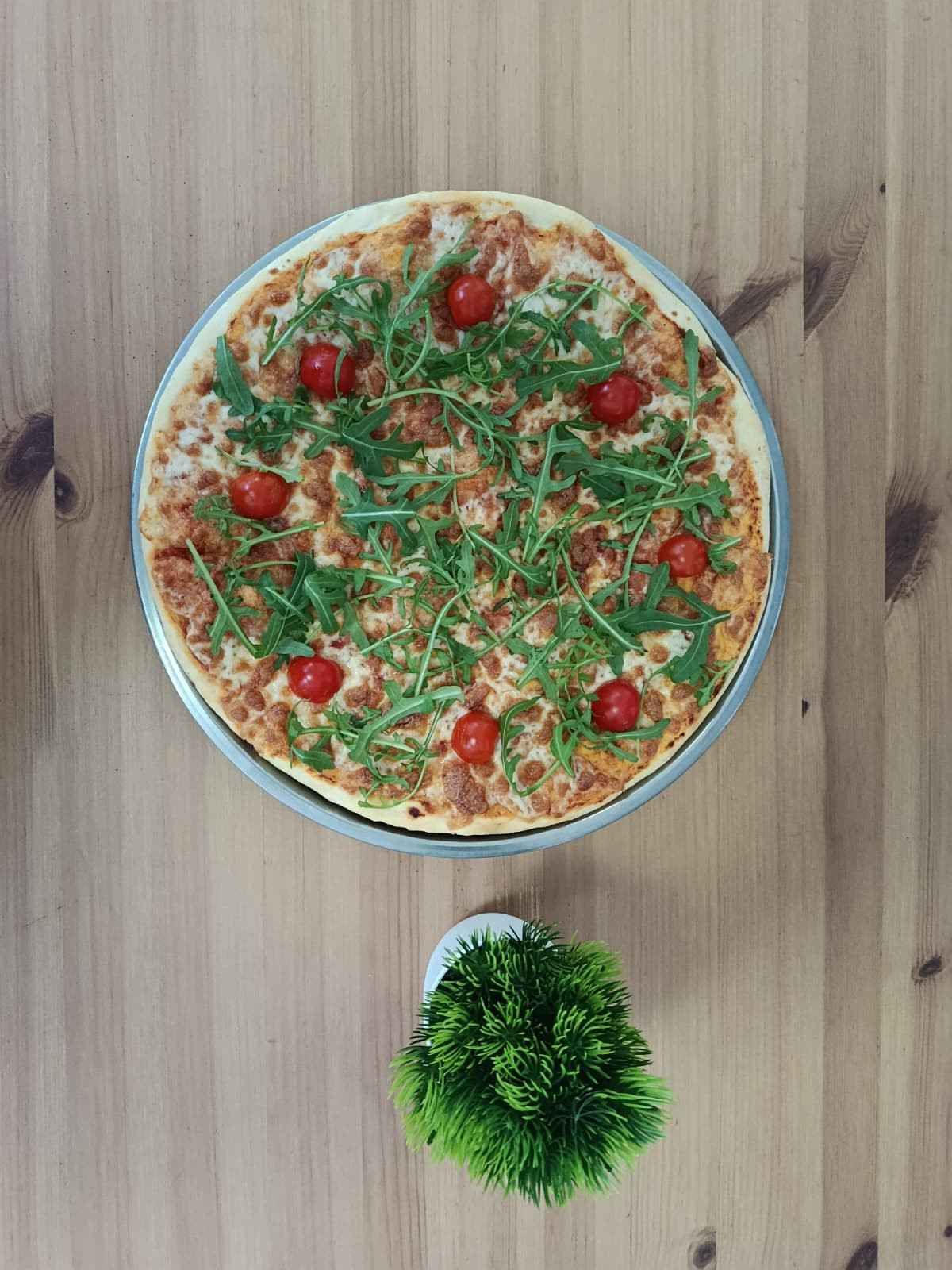 Una pizza de masa fina con rúcula y tomates cherry sobre una mesa de madera, junto a una pequeña planta artificial.